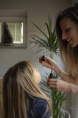 Huzurlu bir yuva ortamında bir kadın, başını arkaya eğen bir çocuğa nazikçe sıvı damlatıcıyı yönetir. Bir ev bitkisi ve yumuşak ışıklandırma dingin ve besleyici çevreye katkı sağlar..