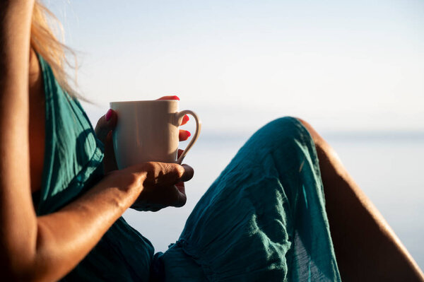 A person relaxes, holding a coffee cup, bathed in warm sunlight. The serene setting and peaceful moment emphasize a quiet escape, dressed in a flowing teal outfit.