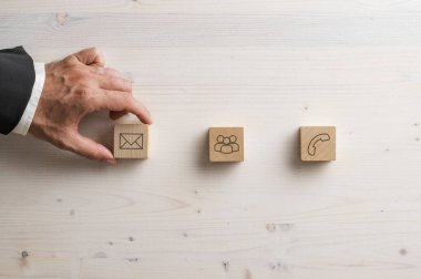 Takım elbiseli bir el, farklı iletişim yöntemlerini sembolize ederek, açık ahşap bir yüzeyde e-posta, grup iletişimi ve telefon temasını temsil eden simgelerle ahşap blokları düzenler..