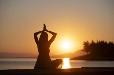 Altın bir günbatımına karşı silueti olan bir insan, sakin bir su kütlesi tarafından meditasyon yapar. Ellerini kaldırarak barışçıl bir jest yapar. Huzur ve dinginliği kucaklar..