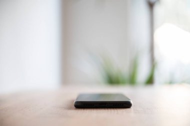 Akıllı telefon, ahşap bir masada düz bir şekilde uzanır, bulanık, ışık dolu bir arkaplanı olan yumuşak bir odak ile yakalanır, sakin, minimalist bir atmosfer yaratır..