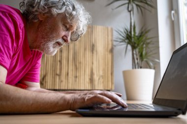 Parlak pembe tişörtlü, odaklanmış yaşlı bir adam, işine dalmış, dizüstü bilgisayarını kullanır. Sahne rahat, bitki dolu bir ev ortamında geçiyor..