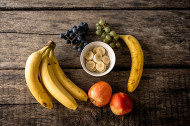 Muz, elma, yeşil ve siyah üzüm gibi çeşitli meyveler ve kırsal ahşap bir yüzeye dizilmiş bir kase dilimlenmiş muz..