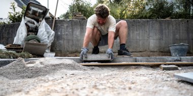 Eldiven ve tişört giyen bir adam kumlu bir zemine kaldırım taşını dikkatlice yerleştirir. Aletler ve inşaat malzemeleriyle çevrili bir yere..