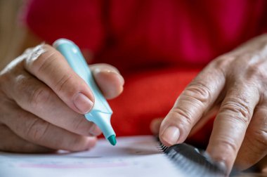 Bir turkuaz fosforlu kalem tutan, bir defterin üzerinde duran, odaklanmayı ve üretkenliği vurgulayan bir elin ayrıntılı yakın çekimi. Kırmızı arkaplan sahneye canlı bir karşıtlık ekler.