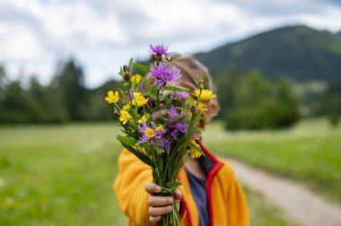 Turuncu ceketli bir çocuk, arka planda yemyeşil manzarası ve tepeleri olan mor ve sarı kır çiçeklerinden oluşan renkli bir buket tutar..