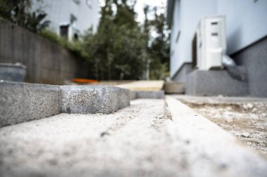 Çağdaş bir konut binasının yanında, kum ve inşaat malzemeleriyle çevrili, kısmen beton bloklar döşenmiş, tamamlanmamış bir kaldırımın yakın görüntüsü.