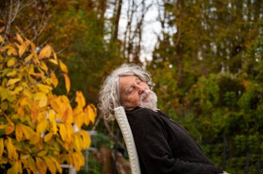 Kıvırcık gri saçlı ve sakallı yaşlı bir adam dışarıda bir sandalyede, gözleri kapalı, sakin bir ormanda canlı sonbahar yapraklarıyla çevrili, dinleniyor..