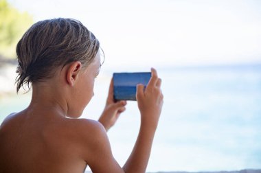 Islak saçlı ve güneş yanığı tenli genç bir çocuk akıllı telefonuyla okyanusun güzel bir deniz kenarı anını korurken fotoğrafını çekiyor..