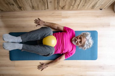 Parlak pembe gömlekli yaşlı bir adam yoga minderinde yatıyor, bacaklarının arasında sarı bir egzersiz topu tutuyor, yüzünde bir gülümsemeyle vücut egzersizi yapıyor..