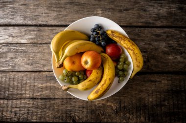 Muz, elma, portakal ve üzümlerle dolu beyaz bir kase, taze ve hafif olgunlaşmış meyvelerin bir karışımını sunan, rustik ahşap bir masada oturur..