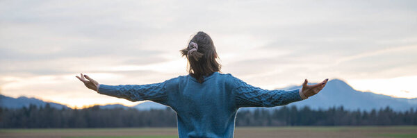 A woman in a blue sweater stands with open arms, facing the mountains and sky at sunset, enjoying a moment of peace and freedom in nature.
