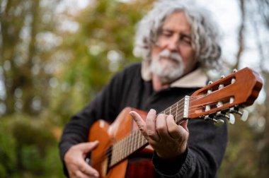 Kıvırcık gri saçlı yaşlı bir adam açık havada akustik gitar çalıyor, müziğe dalmış, bulanık yeşil yapraklarla çevrili..