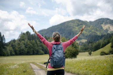 Sırt çantalı bir kadın kollarını kaldırır yemyeşil bir vadide yürürken görkemli dağlarla ve parlak mavi gökyüzüyle..