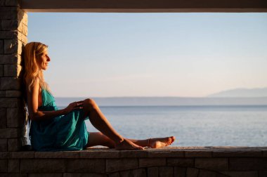 Turkuaz elbiseli bir kadın deniz kenarında dinleniyor, ılık altın bir ışıkla yıkanmış, ufuktaki uzak dağlarla sakin okyanusa bakıyor..
