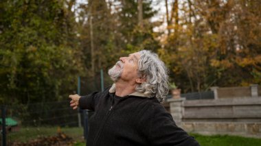 Kıvırcık gri saçlı ve sakallı yaşlı bir adam dışarıda duruyor, kolları açık, huzurlu bir ifadeyle yukarıya bakıyor, sakin bir ortamda sonbahar ağaçlarıyla çevrili..