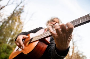 Gri saçlı, akustik gitar çalan, arka planda ağaçlarla gitar tellerine basan yaşlı bir adamın yakın çekimi..