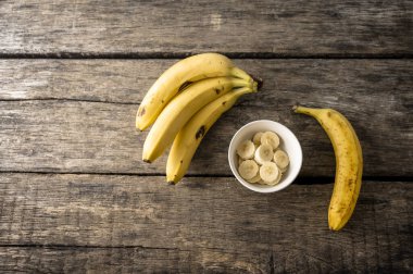  Bir demet olgun muz, tek bir muz, ve küçük beyaz bir kase muz dilimleri, kırsal ahşap bir masa üzerine dizilmiş..