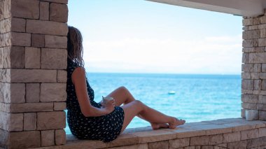 Bir fincan kahveyle rahatlayan bir kadın, taştan bir terastan sakin mavi okyanusa bakıyor..