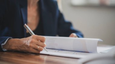 Spor ceketli bir kadın mali belgeleri, sözleşmeleri ve çizelgeleri inceliyor, toplantı sırasında veya çalışma oturumunda not alıyor..