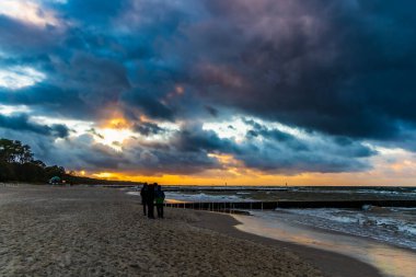 Kumlu kumsalda yürüyen insanlar karanlık bulutlu günbatımında Baltık kıyılarının yanında