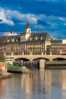 Wroclaw, Polonya - 15 Nisan 2023: Wroclaw Üniversitesi 'nin Odra nehrinin yanındaki güzel binanın cephesi küçük ağaçların ve çalıların arkasından görülüyor