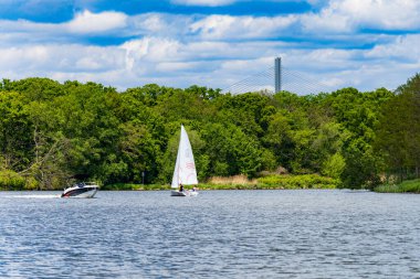 Bulutlu bir günde, yeşil ağaçların ve çalıların önünde akmakta olan iki yüzen kayıkla dolu güzel bir manzara.