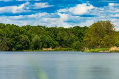 Bulutlu bir günde güzel bir manzara. Yeşil ağaçların ve çalıların önünde akan bir nehir. Üzerinde yüksek bir köprü var.