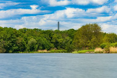 Bulutlu bir günde güzel bir manzara. Yeşil ağaçların ve çalıların önünde akan bir nehir. Üzerinde yüksek bir köprü var.