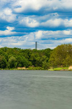 Bulutlu bir günde güzel bir manzara. Yeşil ağaçların ve çalıların önünde akan bir nehir. Üzerinde yüksek bir köprü var.