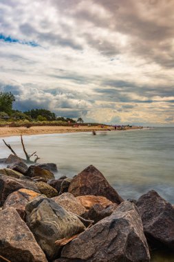 Büyük plajlı, bulutlu, güzel bir manzara. Yürüyen insanlar suyun yanındaki dev kayaların arkasından görülüyorlar.