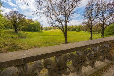 Yeşil Glades 'li büyük yeşil parkın güzel manzarası, bulutlu güneşli bir sabahta ağaçlar küçük bir köprüden parktaki beton çitle görülüyor.