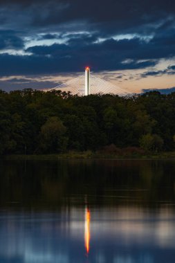 Wroclaw, Polonya - 11 Mayıs 2024: Gökyüzü bulutlu ve yıldızlı gecede sessiz Odra nehrine yansıyan Redzinski köprüsü ile yüksek ağaç ve çalıların güzel manzarası