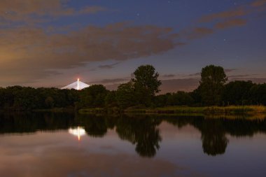 Wroclaw, Poland - May 11 2024: Beautiful nightscape of high trees and bushes with Redzinski bridge behind reflecting in silent Odra river at cloudy and starry night clipart