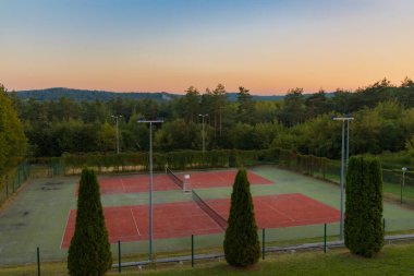 Boş tenis kortları gün batımının ılık renklerinde güneşlenir, yemyeşil bir ortamda yuva yapar ve berrak bir gökyüzünün altında yuvarlanan tepeler.