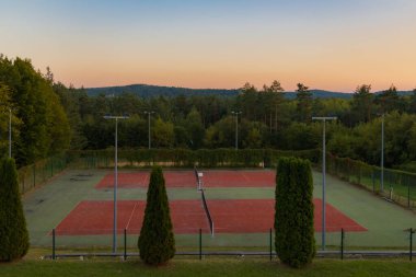 Boş tenis kortları gün batımının ılık renklerinde güneşlenir, yemyeşil bir ortamda yuva yapar ve berrak bir gökyüzünün altında yuvarlanan tepeler.