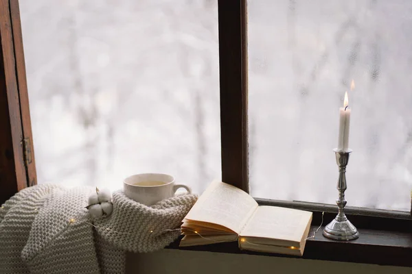 Cozy winter still life. Cup of hot tea and an open book with a warm sweater on a vintage wooden windowsill. Cozy home concept. Sweet home.