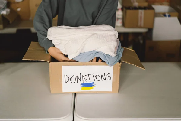 Volunteer teengirl preparing donation boxes for people in need in Ukraine. Donation clothing for refugees from Ukraine, support of war victims, a box with the Ukrainian flag.Humanitarian aid concept.