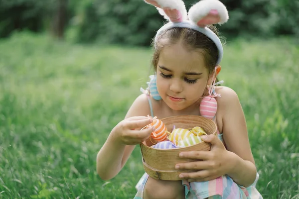Easter egg hunt in spring garden. Funny girl with eggs basket and bunny ears on Easter egg hunt in garden. Children celebrating Easter. Happy easter card