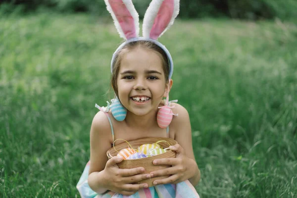 Easter egg hunt in spring garden. Funny girl with eggs basket and bunny ears on Easter egg hunt in garden. Children celebrating Easter. Happy easter card