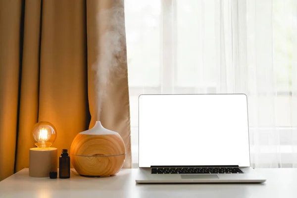 Aroma oil diffuser in work place, laptop and home decor on the table against window.