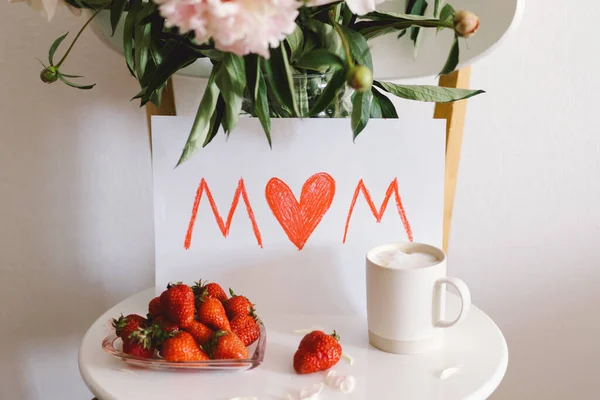 Breakfast for Mothers Day. Heart shaped white plate with fresh strawberries, cup of coffee, gift and Peonys bouquet with gift in bed. Still life composition. Happy Mothers Day.