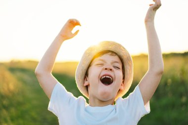 Beyaz tişörtlü gülümseyen küçük bir çocuğun portresi. Gün batımında elini kaldırıyor, oynuyor ve sahada eğleniyor. Mutlu çocuk, yaşam tarzı. Çocuklar için ürünler.