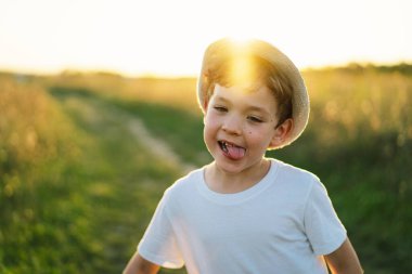 Gün batımında dışarıda oynayan beyaz tişörtlü ve şapkalı gülümseyen küçük bir çocuğun portresi. Mutlu çocuk, yaşam tarzı. Çocuklar için ürünler.