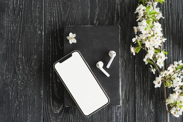 Mockup mobile phone with white blank screen. Book, phone, white wireless bluetooth headphones on rustic wooden table. Audio technology apps, music podcasts books