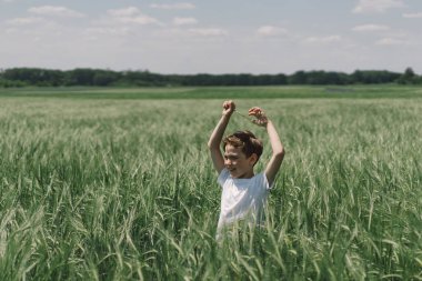 Beyaz tişörtlü ve yeşil arpa tarlasında oynayan gülümseyen bir çocuğun portresi. Mutlu çocuk yaz günü gülüyor ve oynuyor. Doğayı keşfeden çocuk. Meraklı çocuklar için yaz etkinliği