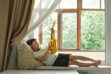 Tatlı çocuk pencerenin yanındaki pencere pervazında sarı Ukulele gitarı çalmayı öğreniyor. Rahat bir ev. Yaz tatili yaşam tarzı.