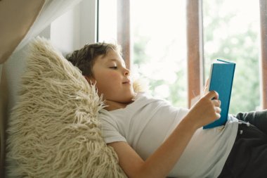 Pencerenin yanındaki pencere pervazında kitap okuyan sevimli çocuk. Güneşli bir günde evde dinlenen bir çocuk. Summer. Ev Okulu ve Eğitim Konsepti