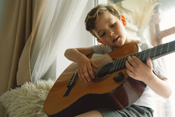 Cute boy learns to play the classical guitar on the windowsill near the window. Cozy home. Summer holidays lifestyle.