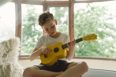 Tatlı çocuk pencerenin yanındaki pencere pervazında sarı Ukulele gitarı çalmayı öğreniyor. Rahat bir ev. Yaz tatili yaşam tarzı.